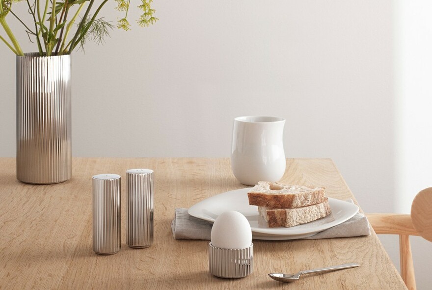 Silver table setting, including vase, salt and pepper shakers and egg cup, on a wooden table.