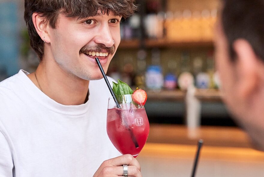 A young man with a moustache, drinking a red cocktail with a straw.