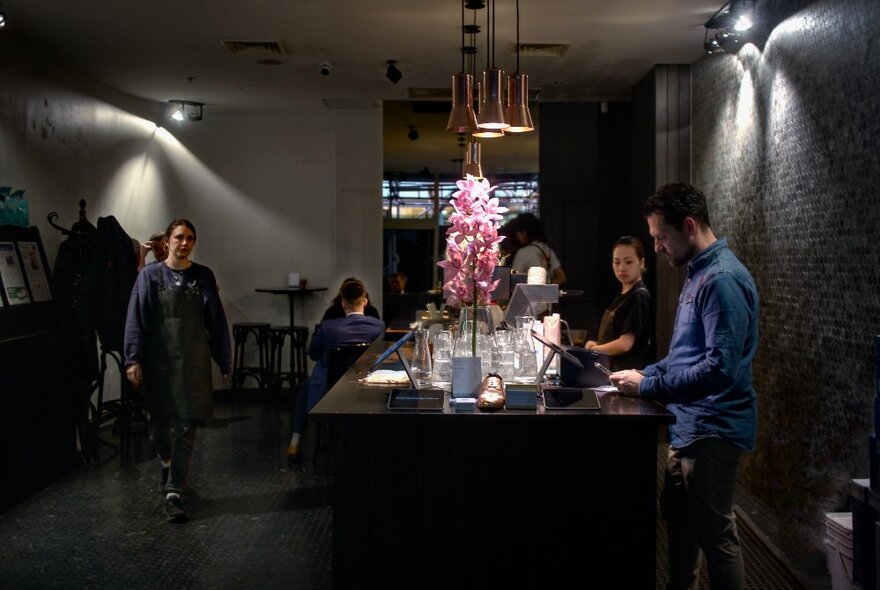 Interior of Rosso Espresso Bar, with staff behind a service counter that is lit by hanging brass lamps, and a person walking through the cafe space.