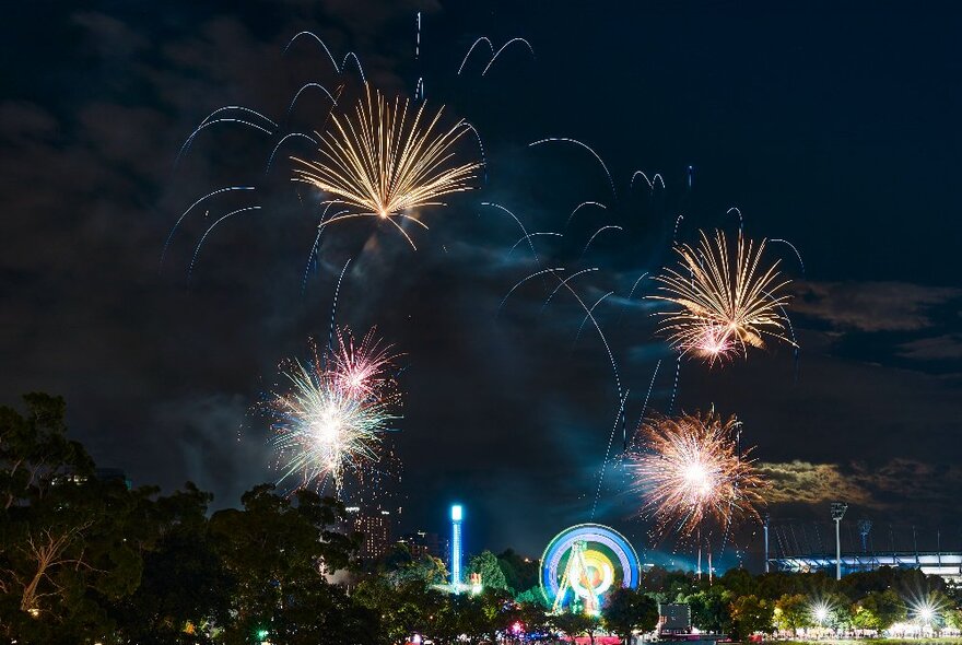 Four golden starburst fireworks against a dark sky, with people and carnival rides just visible underneath.