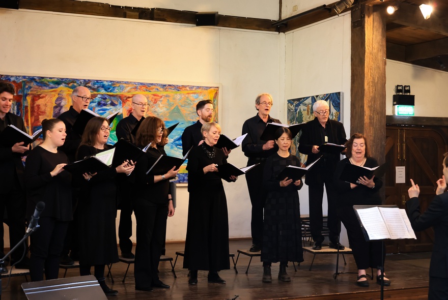 A small choir all dressed in black singing from open books in a gallery setting. 
