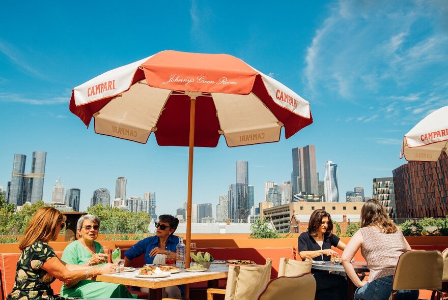 Five women dining under a red umbrella on a sunny city rooftop bar.