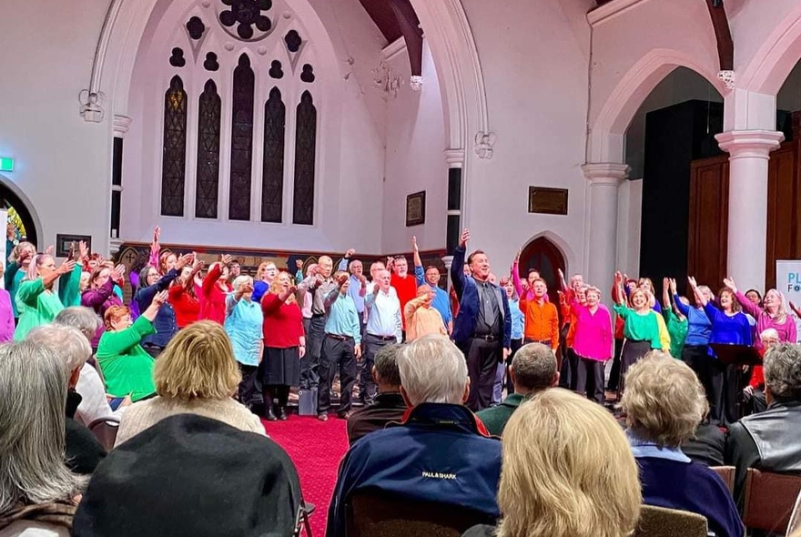 A large choir singing at a church hall, each memeber wearing brightly coloured shirt.