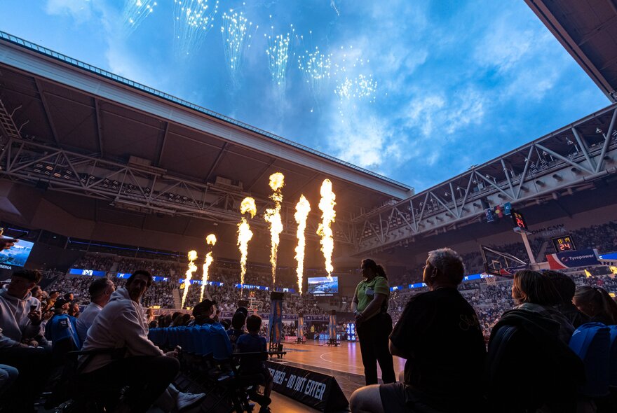 A crowd of basketball fans inside John Cain Arena, the retractable roof is open to a blue sky, and there are flames shooting up into the air from centre court as part of the pre-game entertainment