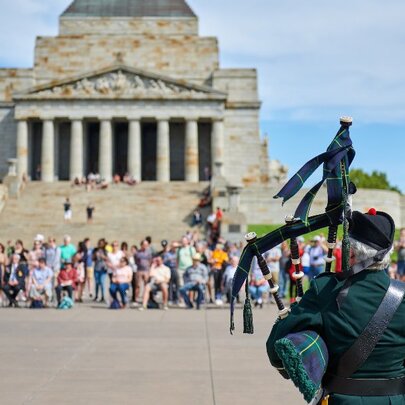 People gathered in front of the Shrine of Remembrance and a man playing bagpipes with his back to the camera.