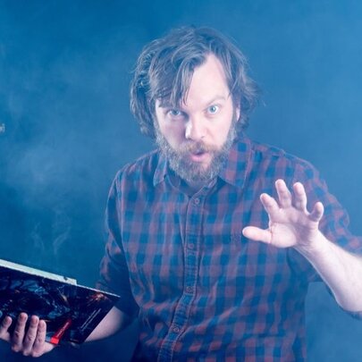 Bearded man holding an open book in a misty space, holding his hand in the air.