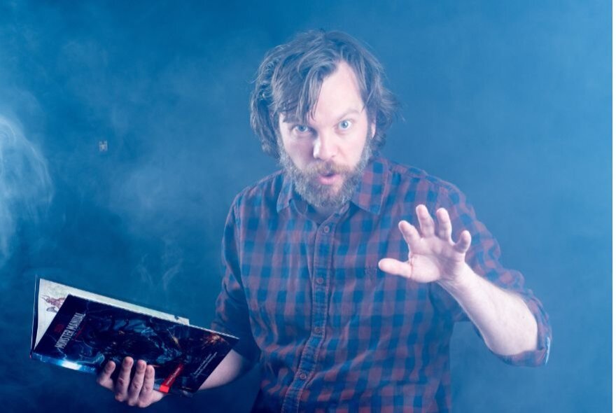 Bearded man holding an open book in a misty space, holding his hand in the air.