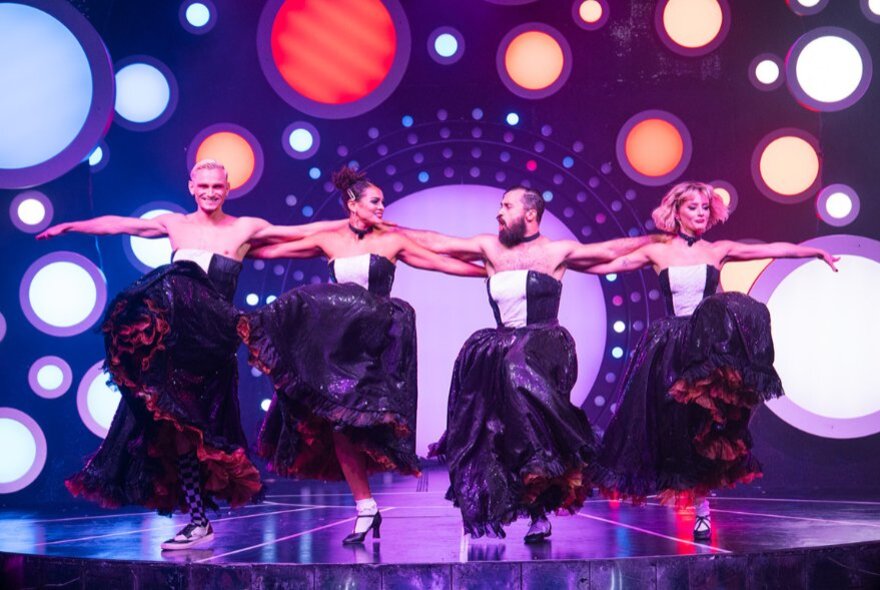 Four people wearing strapless voluminous dresses dancing the Can Can on a cabaret stage.