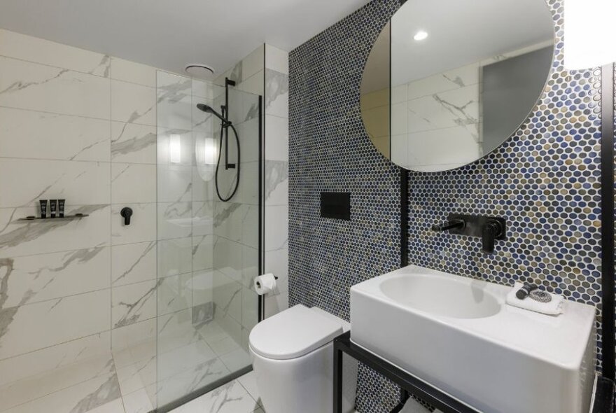 Hotel room bathroom with shower cubicle, white fittings and tiled wall with circular mirror.