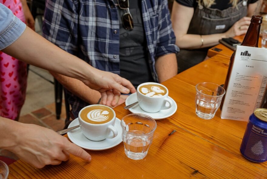 Two white ceramic cups of coffee with latte art being served on saucers at a wooden table in a cafe.