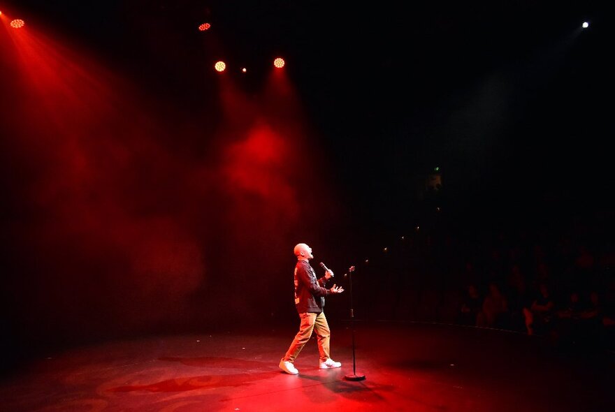 A standup comedian performing solo under a spotlight on a large stage.