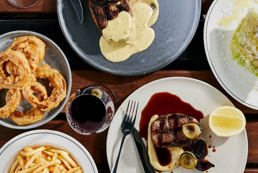 Looking down at plates of food on a dining table that includes steak, chips, onion rings, and a glass of wine. 