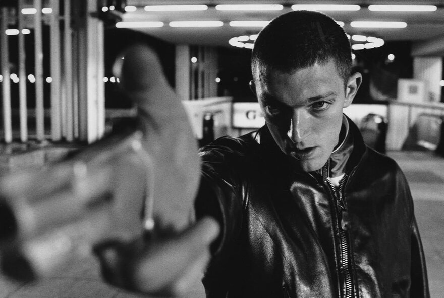 A still-frame from black and white French film, La Haine, depicting a youth wearing a leather jacket and brandishing a gun.