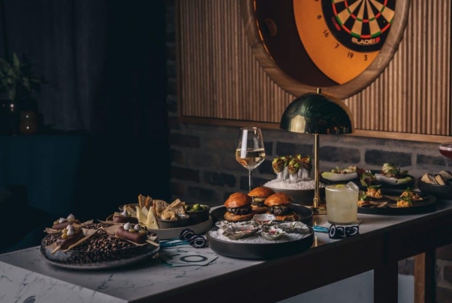 Grey bench laden with foods and drinks; dart board at rear right.