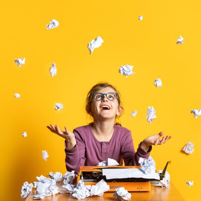 A young teeneager, sitting in front of a old-fashioned typewriter at a desk, surrounded by scrunched up balls of paper on the desk and in the air around her, against a yellow background.
