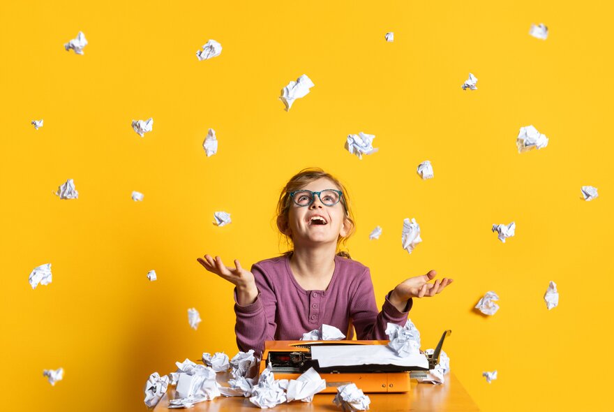 A young teeneager, sitting in front of a old-fashioned typewriter at a desk, surrounded by scrunched up balls of paper on the desk and in the air around her, against a yellow background.