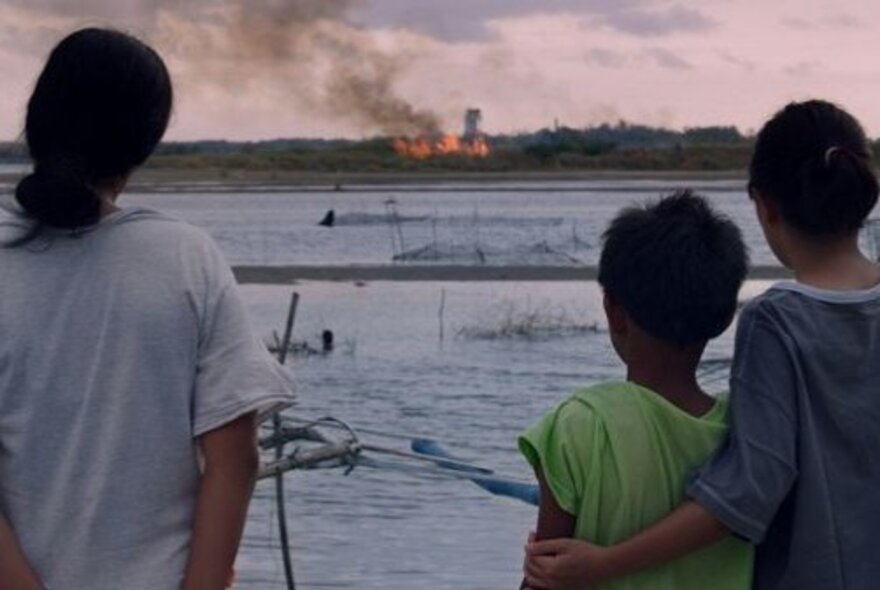 Still from a film of three people viewed from behind, standing on a shore or dock, looking out across a body of water towards a distant fire or explosion on the opposite bank.