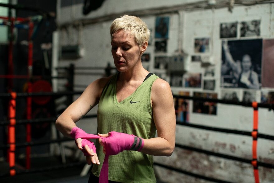 A woman wearing a green singlet wrapping in her hands in pink tape in a boxing ring at a gym.