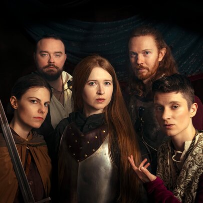 Actors dressed in medieval costumes posing grouped together with serious expressions. 