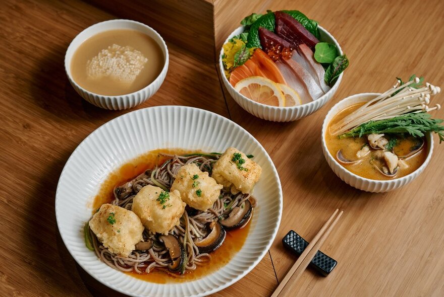 A bowl of dumplings in broth, with smaller side dishes of food, on a pale timber table surface.