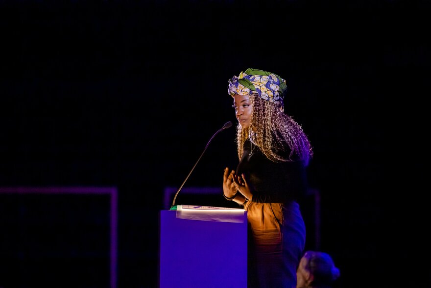 A woman wearing a floral headscarf, reading at a podium.