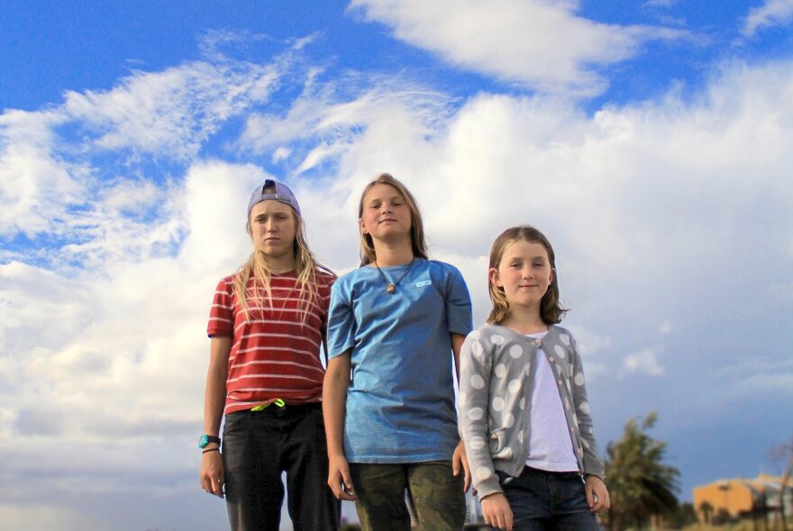 Three young, female Olympic skateboarding hopefuls, with blue sky and clouds behind them.