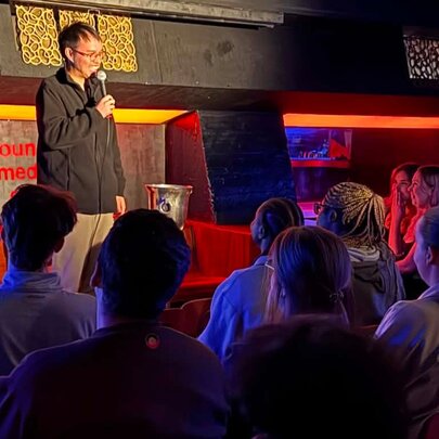 Stand-up comedian standing on a stage holding a microphone in front of a small seated audience.