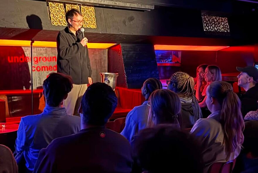 Stand-up comedian standing on a stage holding a microphone in front of a small seated audience.