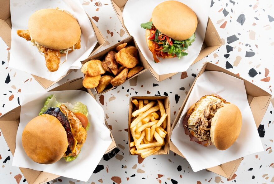 Overhead view of four burgers in takeaway cardboard containers, with fries, on a patterned table top.