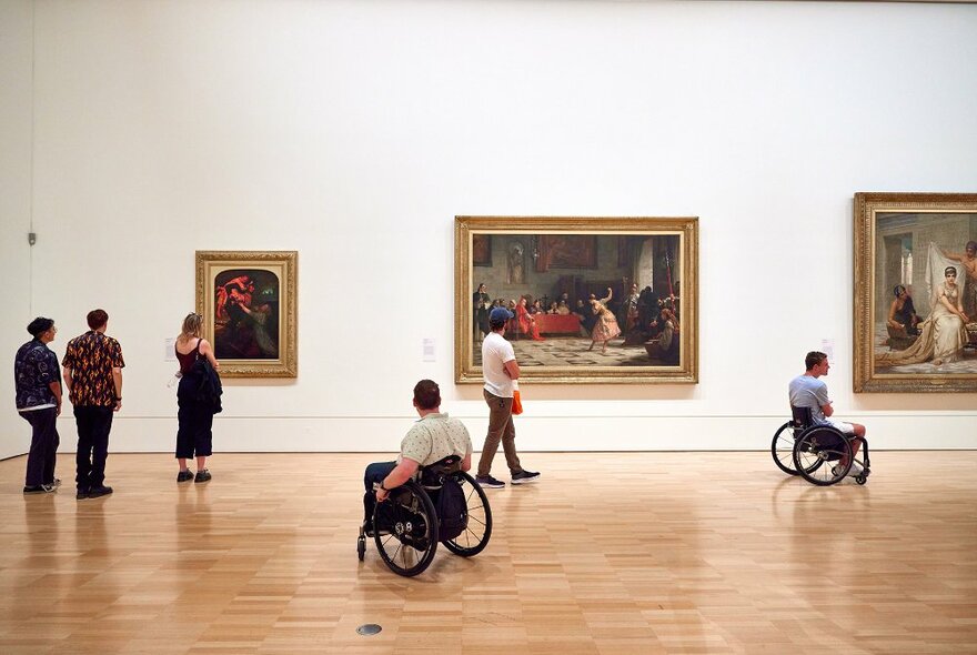 People looking at paintings in a spacious gallery. Two people are using wheelchairs.