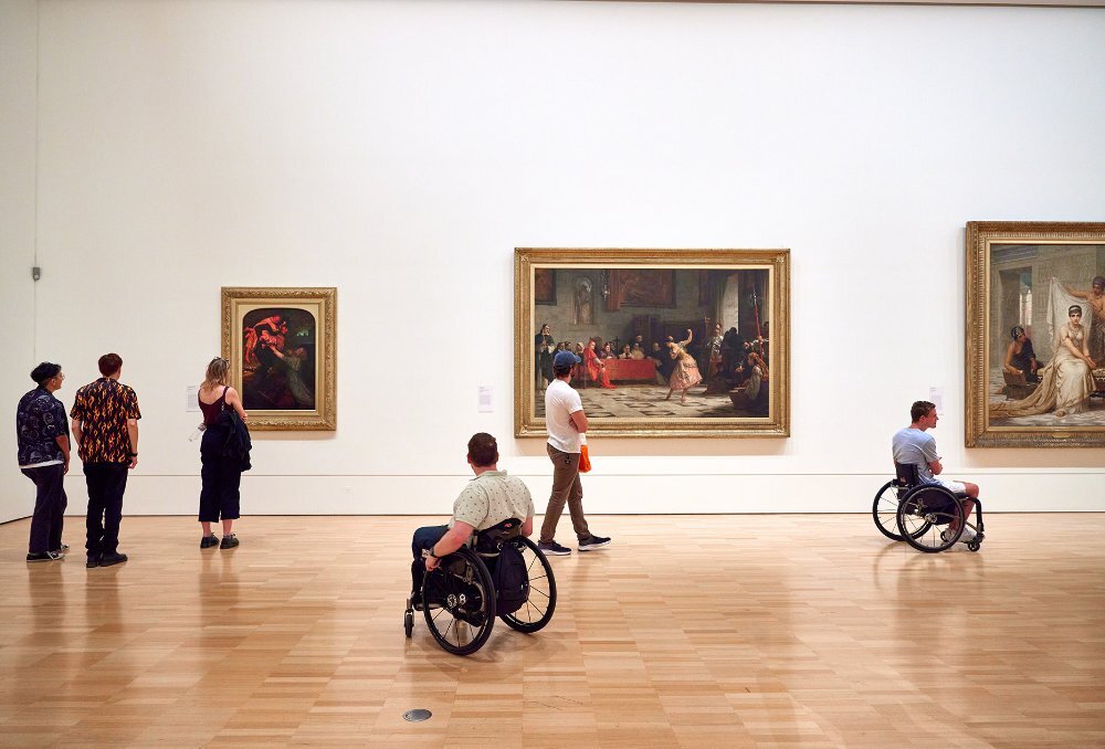 People looking at paintings in a spacious gallery. Two people are using wheelchairs.