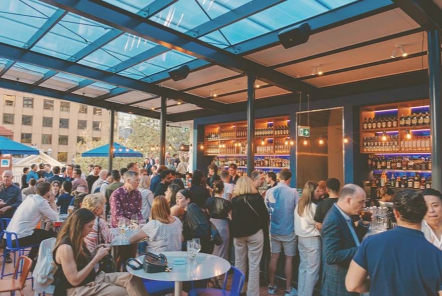 An outdoor rooftop bar, with people socialising and drinking, a large bar in the rear of the space, with views of Melbourne buildings in the background.