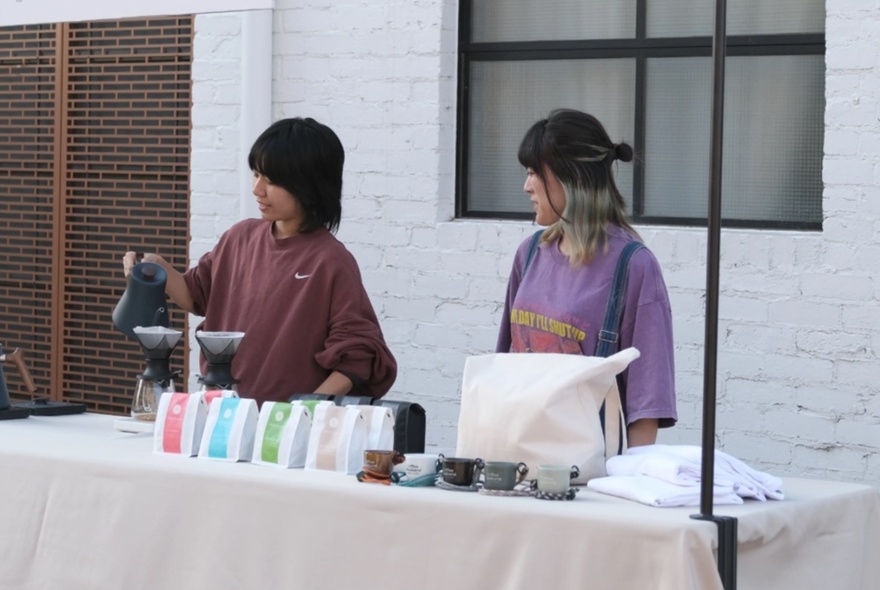 Two people behind a table at a stall, one of them making a filter coffee, with bags of coffee at the front of the table.