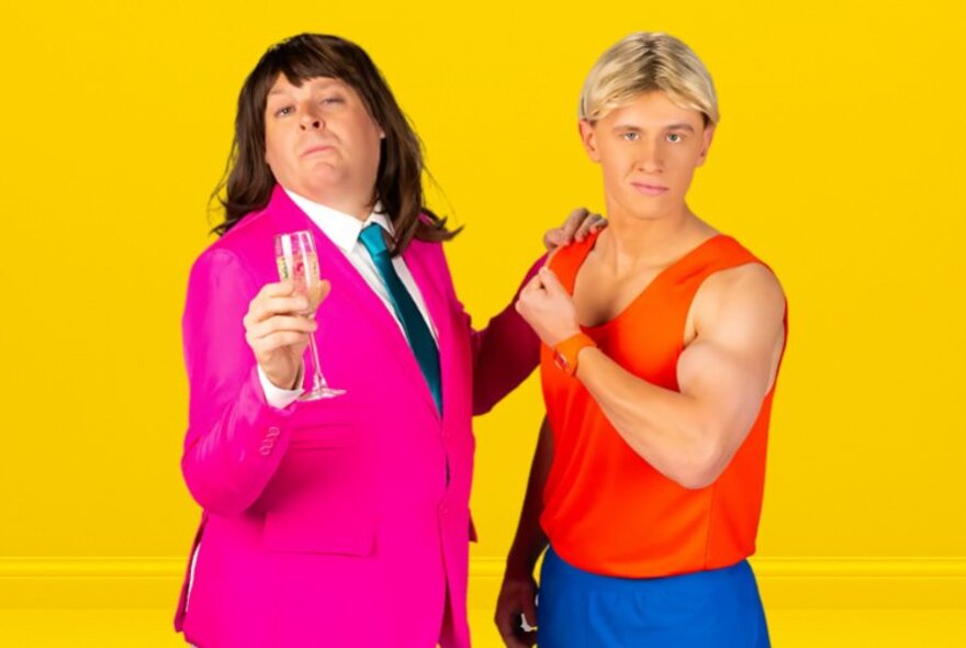 Actor with long black wig and pink jacket holding a glass of bubbles with a young man with blond wig wearing an orange singlet and tracksuit pants.