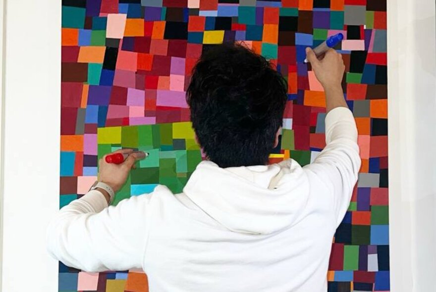 Artist colouring in squares of colour on an artwork on a wall.