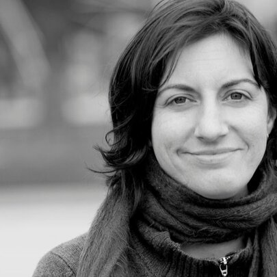 A black and white close-up of a smiling woman with shoulder-length dark hair, wearing a thick, dark scarf wrapped around her neck and a zippered jacket.