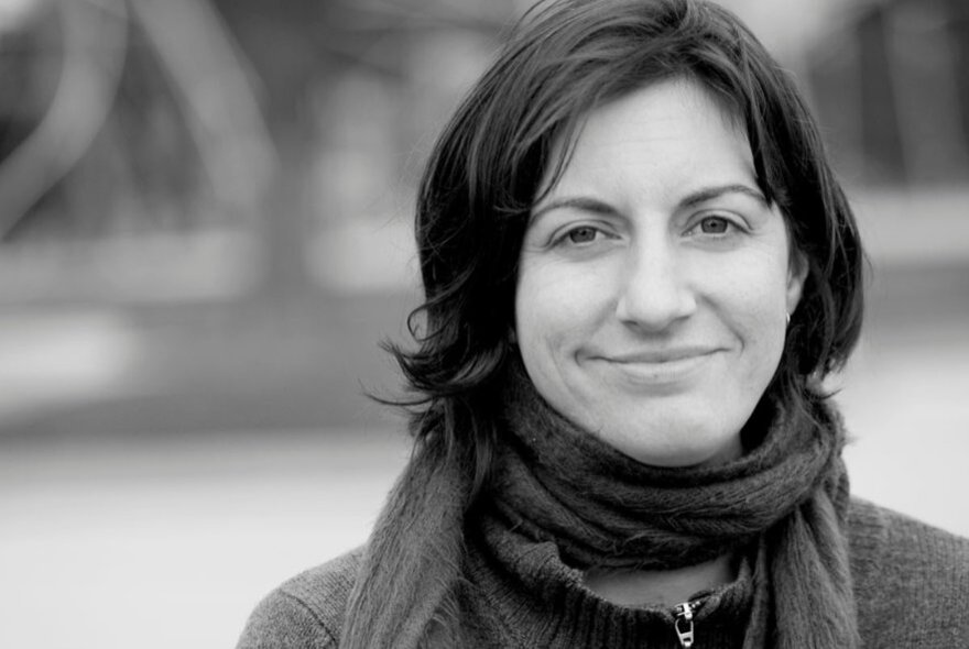 A black and white close-up of a smiling woman with shoulder-length dark hair, wearing a thick, dark scarf wrapped around her neck and a zippered jacket.