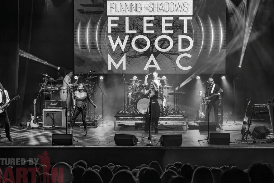 A band of musicians and singers performing live on stage, with a sign on the screen behind them that reads FLEET WOOD MAC; black and white image.