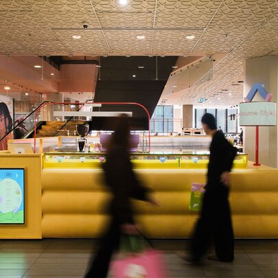 A yellow pop up shop in the centre of a shopping mall, with two people in black blurred as they pass by.