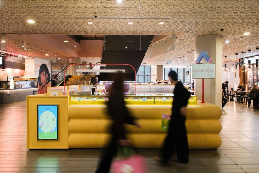 A yellow pop up shop in the centre of a shopping mall, with two people in black blurred as they pass by.