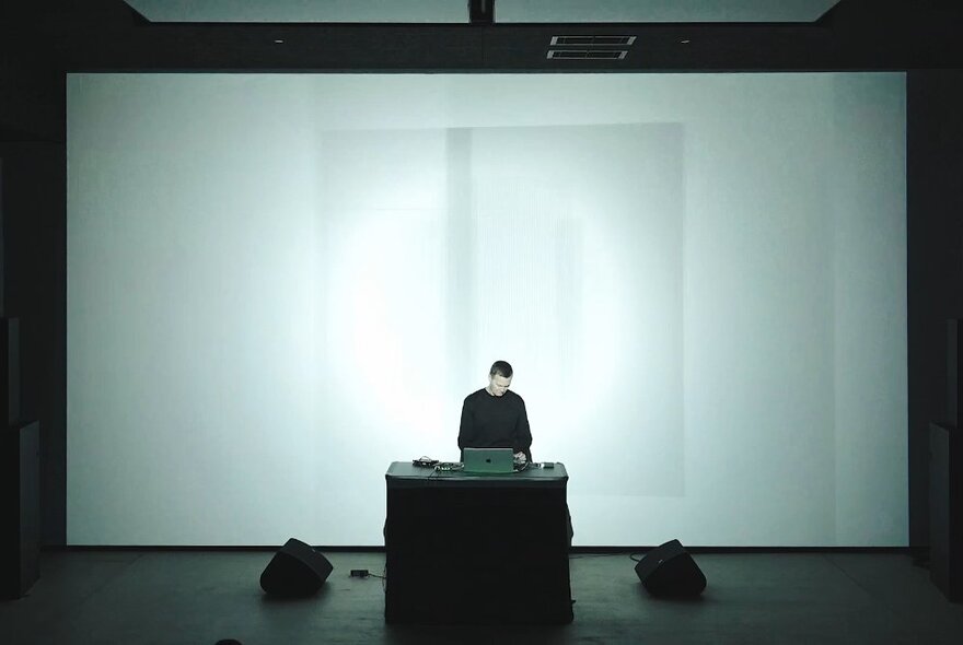 Person on a stage, using a laptop that rests on a small table in front of him, behind him is a large cinema sized screen.