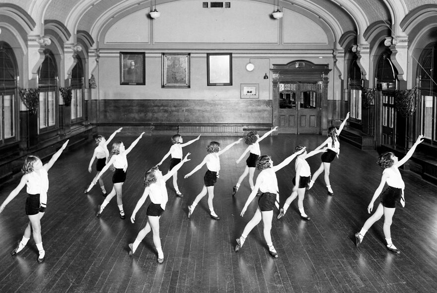 Vintage black and white photograph of a dance class taking place inside the ballroom on the third floor of Melbourne's Flinders Street Station, shows a group of young people in white tops and black shorts performing a dance routine in a large hall with a wooden floor and arched ceiling.