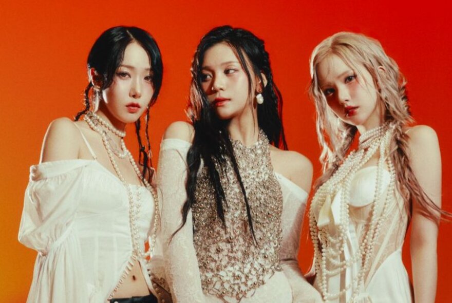 Three women all in different cream outfits, pose for a band photo with serious expressions against an orange/red background.
