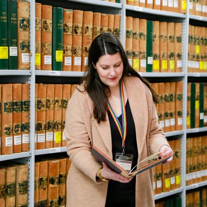 A woman looking at an old archival book with yellow and green binding in front of packed shelves of the same kind of book.