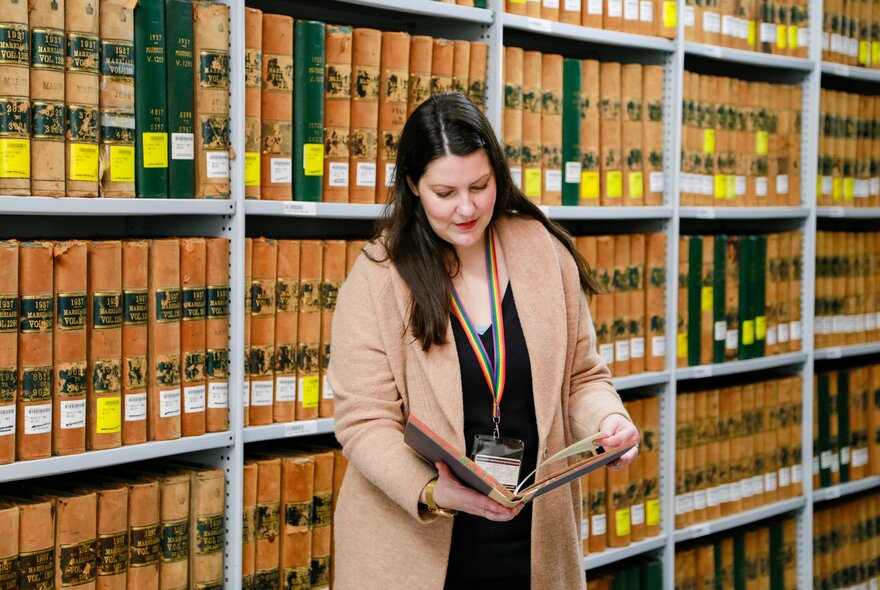 A woman looking at an old archival book with yellow and green binding in front of packed shelves of the same kind of book.