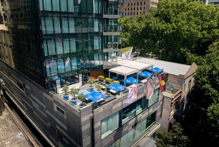 Aerial, high-angle view of a modern glass hotel building featuring an outdoor rooftop terrace with lounge seating under umbrellas, and lush green trees bordering the side.