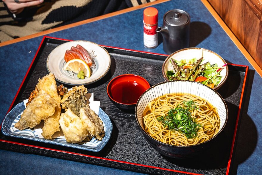 A tray on a dining table with a plate of tempura, a bowl of noodles in broth, side dishes and condiments.
