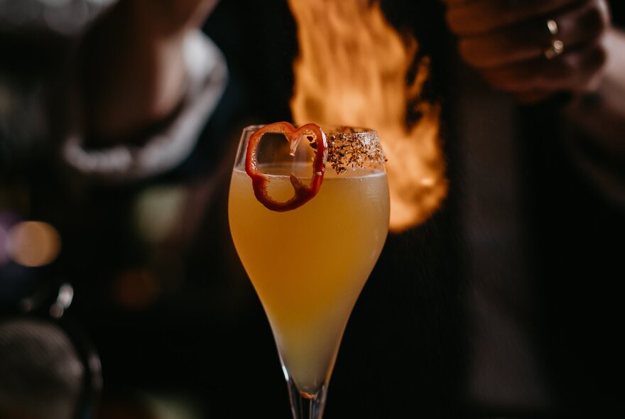 A cocktail with a garnish and fire visible behind the glass. 