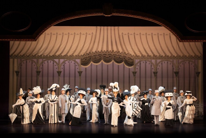 A scene from the musical My Fair Lady, with a large ensemble cast on stage for the Ascot scene, in black and white attire. A scene from the musical My Fair Lady, with a large ensemble cast singing and dancing on stage.
