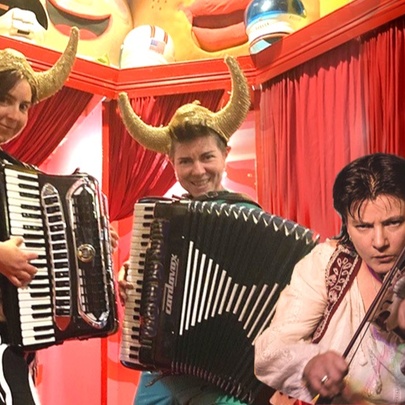 Two accordionists and a violinst wearing cow horns, performing a red-curtained tent.